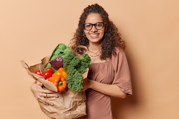 Horizontal shot of pleasant looking young woman with dark curly hair holds big grocery bag full of...