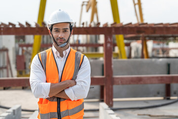 Portrait of male civil engineer wearing safety vest with white helmet standing arms crossed at factory making precast concrete wall for real estate housing.