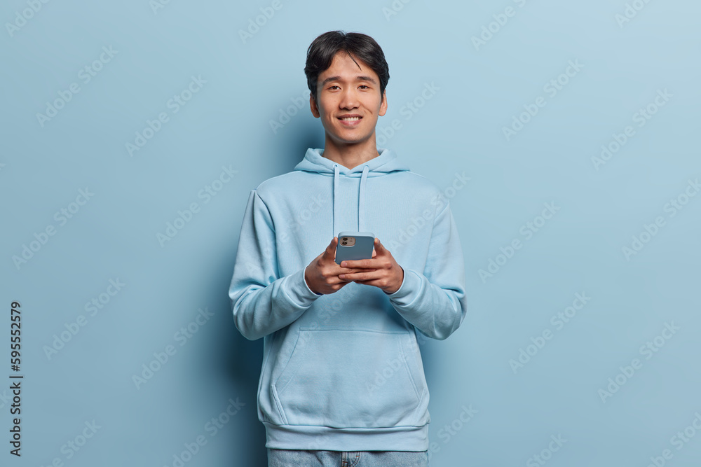 Wall mural People and modern technologies concept. Handsome dark haired Chinese man smiles gently holds cellphone makes shopping online text sms message dressed in casual hoodie isolated over blue background - Wall murals