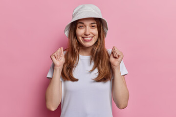 Fototapeta na wymiar Cheerful woman with happy expression smiles broadly clenches fists and rejoices over something wears panama and casual white t shirt isolated over pink background anticipates for news announcement