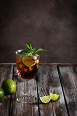 Cuba Libre in a glass with brown rum, cola lime, mint on a dark wooden background. Side view, selective focus.