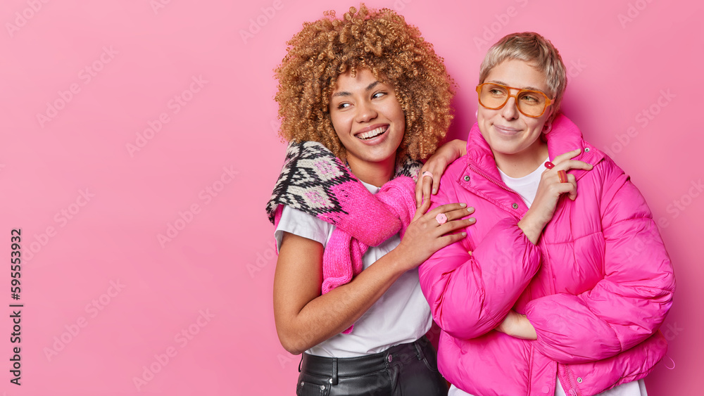 Wall mural Two friendly women dressed in fashionable clothes stand closely to each other focused aside with interest spend free time together isolated over pink background copy space on left side for your promo - Wall murals