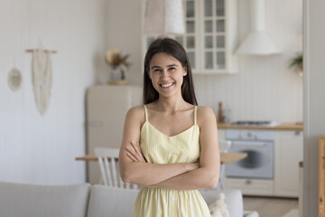 Naklejka premium Positive confident pretty brunette young adult Latin girl wearing sleeveless summer dress, looking at camera with crossed hands, smiling, posing at home. Indoor female portrait
