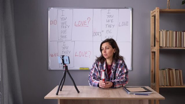 Indian female teacher pointing at whiteboard, explaining English grammar rules to adult students. Tutor recording video lesson using smartphone on tripod. E-learning Concept.