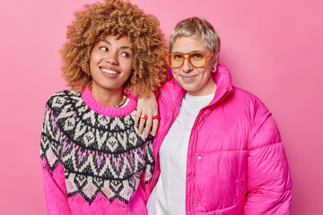 Studio shot of cheerful young European women meet together wear jumper and jacket stand next to each other feel happy isolated over pink background. Fashionable female models pose in studio.