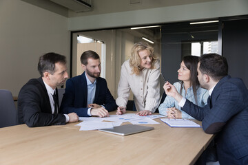 Positive mature marketing coach, business mentor woman discussing paper reports with employees, presenting statistic data, explaining charts, speaking to colleagues over documents at meeting table