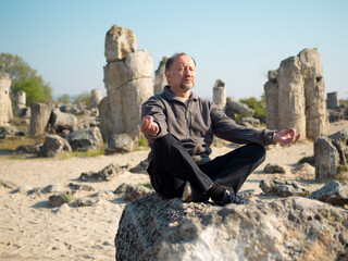 Outdoor Senior man meditating in lotus pose. Sunrise meditation portrait in a magical place Stone Forest similar to Stonehenge, mindfulness concept