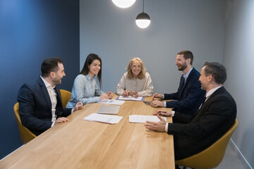 Positive happy coworkers having fun at table, talking, laughing, smiling, discussing new ideas for project, joking. Senior female group leader holding corporate team meeting in small office