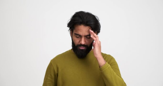 Exhausted bearded young man removing eyeglasses and rubbing head in pain while standing isolated against white background