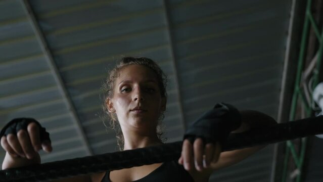 Woman boxer hung from ring ropes in gym after fight. Female muay thai fighter covered in sweat and wearing black sportswear and bandages rests and breathes while looking at camera
