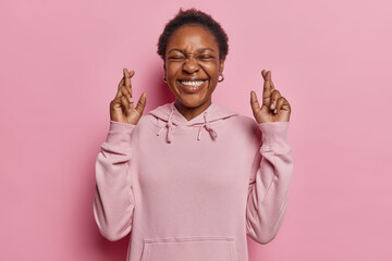 I have to win. Positive dark skinned woman with broad smile and closed eyes raises fingers crossed makes desirable wish waits for good news has white even teeth wears hoodie isolated over pink wall - obrazy, fototapety, plakaty