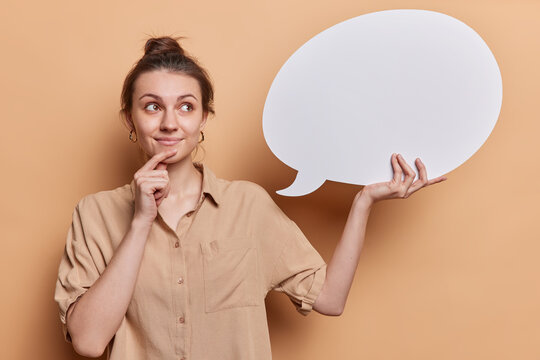 Information idea and thought concept. Pensive European woman with hair bun holds empty speach bubble keeps hand on chin dressed in shirt isolated over beige background. Let me think about it
