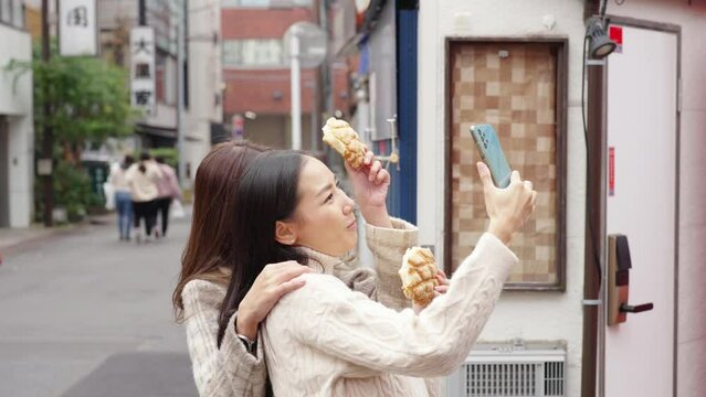 4K Asian woman friends using mobile phone taking selfie together during eating sweet bread at Asakusa district, Tokyo, Japan. Happy girl enjoy outdoor lifestyle travel city street on holiday vacation.