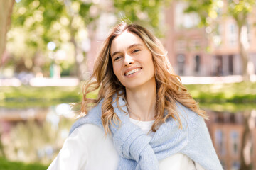 Beautiful woman smiling and look at camera at park at sunny day. Outdoor portrait of a smiling curly blonde girl. Happy cheerful girl laughing at park wear blue sweater, white longsleeves.
