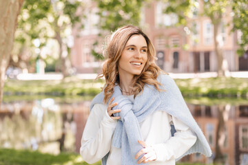 Obraz premium Beautiful woman smiling and look at camera at park at sunny day. Outdoor portrait of a smiling curly blonde girl. Happy cheerful girl laughing at park, look at side, touch sweater, show toothy smile.
