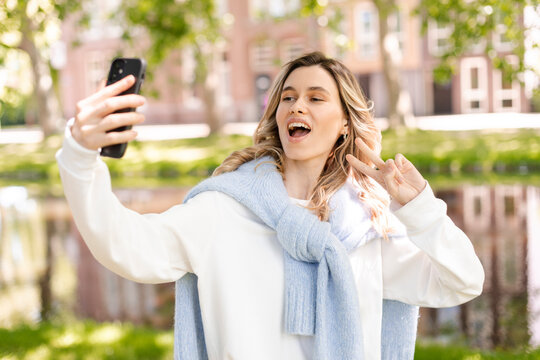 Image of smiling young curly hair blonde woman taking selfie photo holding her phone while walking in park near river. Girl make peace gesture, show two fingers, look happy and smiling.