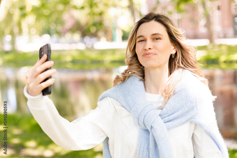 Wall mural Image of smiling young curly hair blonde woman taking selfie photo holding her phone while walking in park near river. Girl make video call using her phone, look happy and smiling.