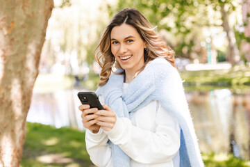 Young stylish curly blonde woman writes message on smartphone walking on park near river, hipster tourist traveling in town and planning trip using mobile phone. Girl chatting on mobile and smiling.