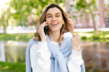 Young happy blonde woman with curly hair received good unexpected news on the phone call in the park. Work promotion, Job offer, great news, girl make win gesture wear blue sweater, white t-shirt.