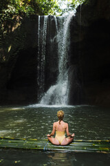 A young girl in a yellow swimsuit meditates near a waterfall.