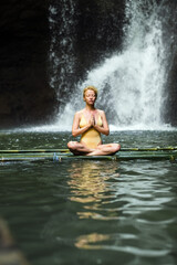 Fototapeta na wymiar The girl is meditating, her hands are folded in namaste, in the jungle against the backdrop of a waterfall, dressed in a yellow swimsuit.