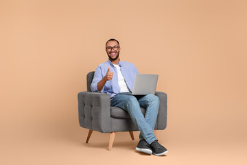 Smiling young man with laptop sitting in armchair and showing thumbs up on beige background - Powered by Adobe