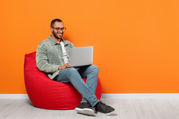 Smiling young man working with laptop on beanbag chair near orange wall, space for text