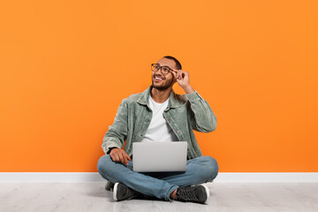 Smiling young man with laptop sitting on floor near orange wall, space for text - Powered by Adobe