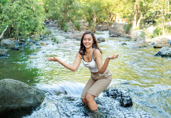 Asian woman in a summer dress splashes in the water at a waterfall cascading down a mountainside. The sun shines brightly overhead, and the woman's laughter echoes through the air.
