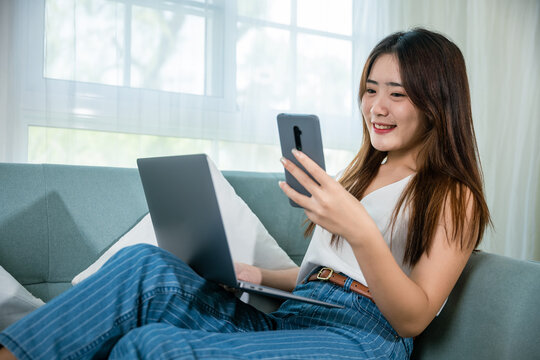 Asian young woman using smart mobile phone while sitting on sofa in living room at home with laptop computer, Happy female use netbook browsing and payment shopping online with smartphone