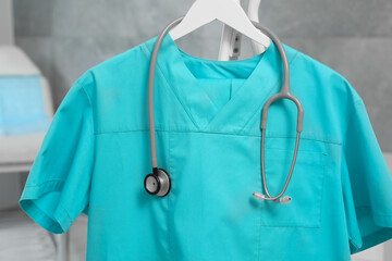 Turquoise medical uniform and stethoscope on rack in clinic, closeup