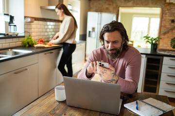 Young adult man using a smart phone while his wife is working in the kitchen behind him