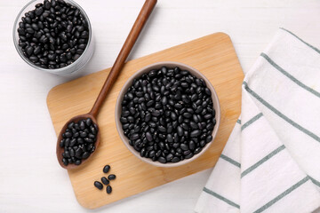 Fototapeta na wymiar Bowl and spoon of raw black beans on white wooden table, flat lay