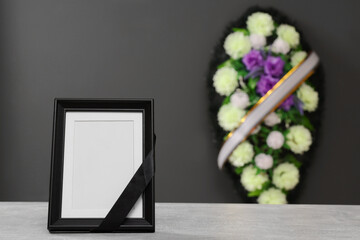 Photo frame with black ribbon on light table and wreath of flowers near grey wall indoors, space...