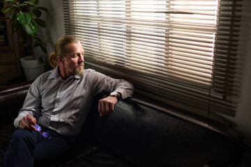 Aged male psychologist in shirt and pants sitting on leather couch by window and looking through venetian blinds while having rest