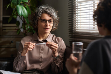 Confident mature female psychologist in eyeglasses looking at patient while listening to description of her problems during consultation