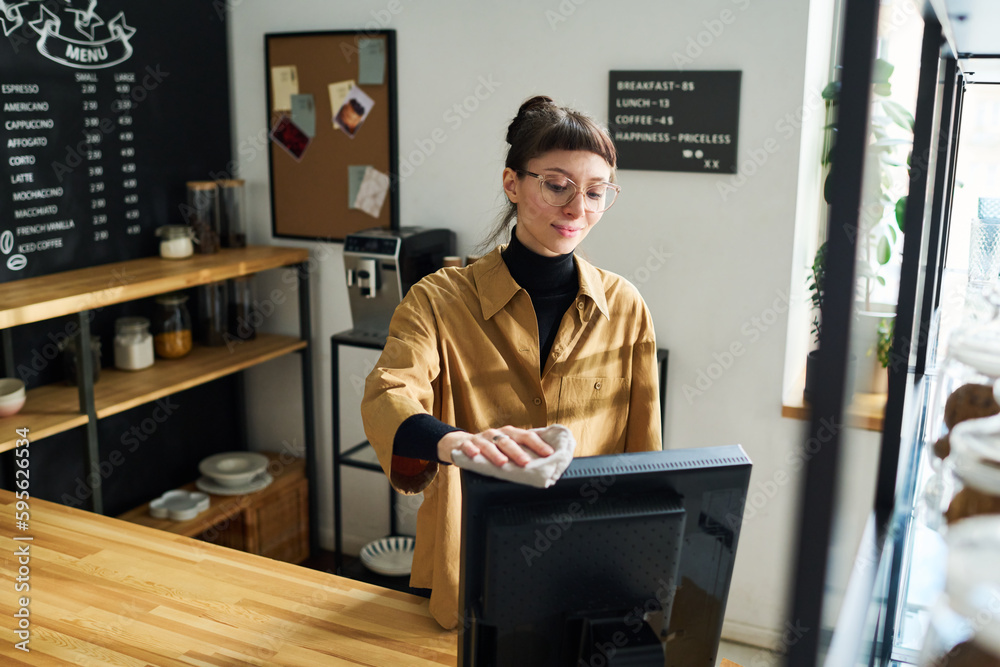 Poster young female clerk with duster wiping computer monitor while standing by workplace against boards an