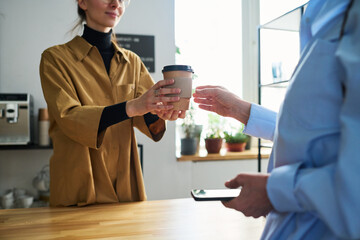 Close-up of young cafe clerk in uniform passing two cups of coffee to mature female guest over counter desk while serving her