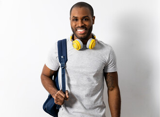 A dark-skinned man poses on a white background with headphones and a backpack. The man has a happy,...