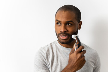 Portrait of a dark-skinned man is using an electric razor to shave his beard on a white background....