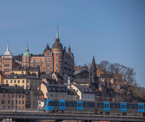 Fototapeta premium Old houses and the house Laurinska Huset with spire on the hill Maria Berget, a tram passing a bridge in front, a sunny spring day in Stockholm