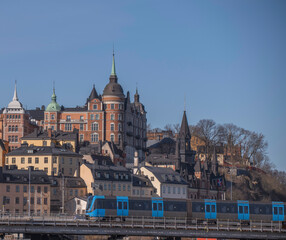 Old houses and the house Laurinska Huset with spire on the hill Maria Berget, a tram passing a bridge in front, a sunny spring day in Stockholm