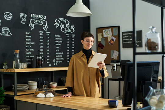 Young smiling clerk in uniform looking through online orders on tablet screen while standing by counter desk against board with menu