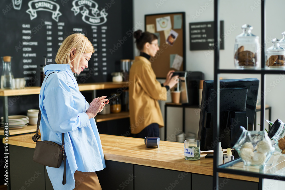Poster side view of mature blond woman texting in smartphone while standing by counter desk and waiting for