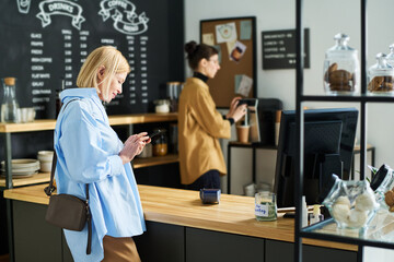 Side view of mature blond woman texting in smartphone while standing by counter desk and waiting for coffee being prepared by clerk