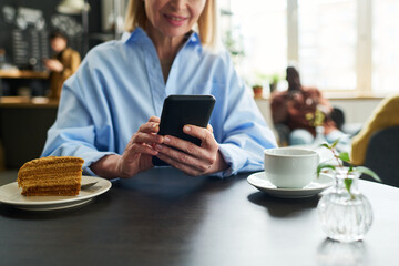 Close-up of mature female consumer using smartphone by table served with cup of tea and piece of cake while having breakfast in cafe