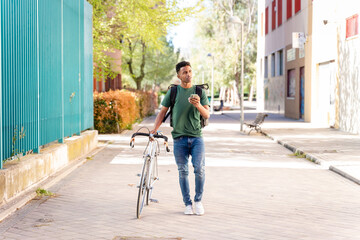 Fototapeta premium Cheerful ethnic man with bicycle walking with backpack delivering food