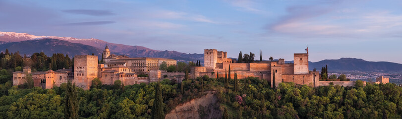 Alhambra is medieval palace and fortress complex with Sierra Nevada snowy mountains in Granada, Andalusia, Spain. Known as a Capital of Nasrid Kingdom or Emirate of Granada. Panoramic sunset hour time - obrazy, fototapety, plakaty