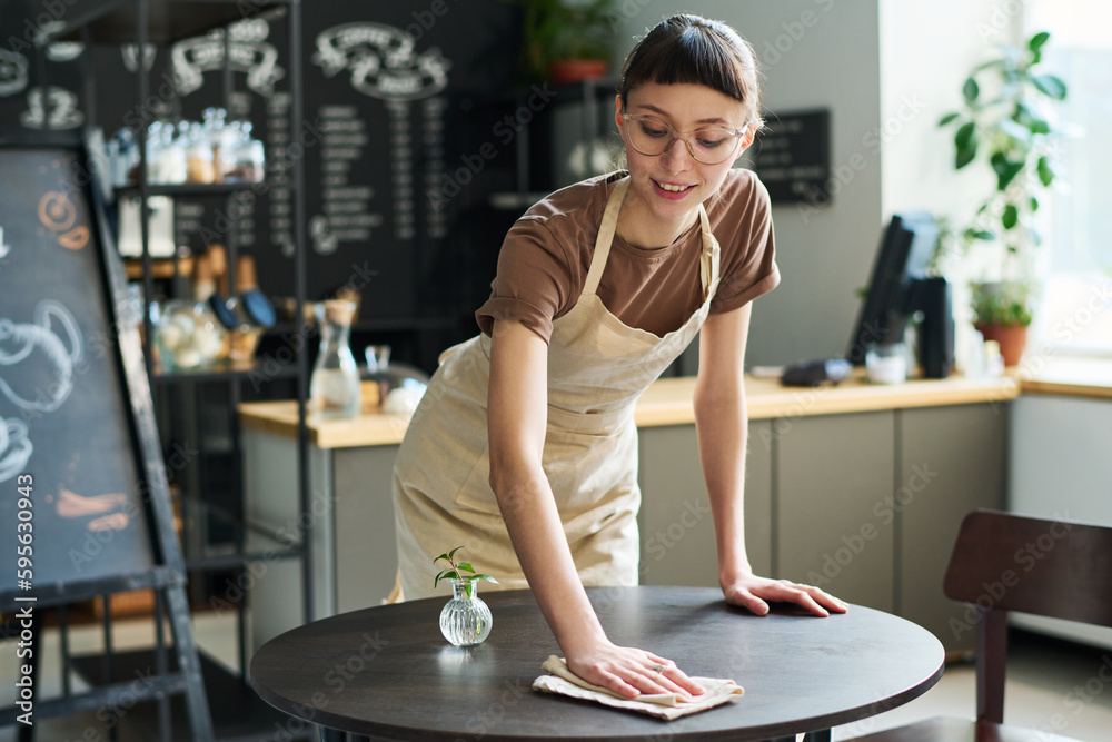 Poster Young smiling waitress in apron bending over small round table and cleaning it with wipe while preparing it for new guests of cafe
