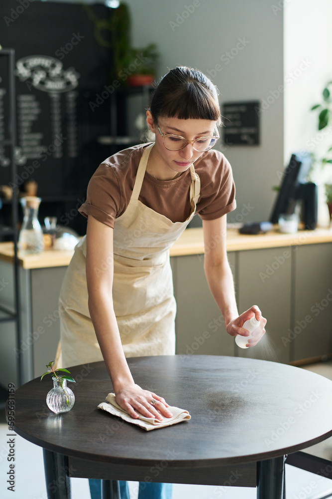 Poster young brunette woman in apron spraying sanitizer on surface of table and cleaning it with wipe while